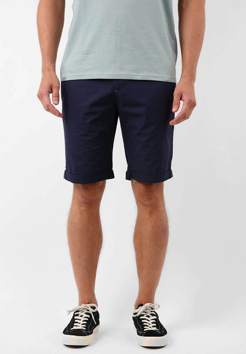 Shorts bleu marine arrivant au genou en coton, avec des revers roulés et des poches latérales, associés à des baskets noires et un t-shirt vert clair.