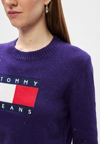 Μωβ πλεκτό πουλόβερ με ριχτό λαιμό, που διαθέτει λογότυπο σε κόκκινο, λευκό και μπλε. Ασημένιες λεπτομέρειες προσθέτουν υφή.