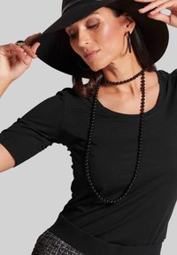 Schwarzes T-Shirt mit kurzen Ärmeln, mehrlagige schwarze Perlenkette und schwarzer Hut mit breitem Rand. Glattes Material, taillierte Passform, neutraler Hintergrund.