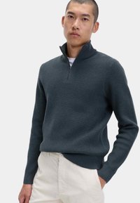 Dunkelgrauer gerippter Pullover mit einem Viertel-Reißverschluss-Kragen, langen Ärmeln und einer strukturierten Oberfläche, getragen über einem weißen T-Shirt und hellen Hosen.