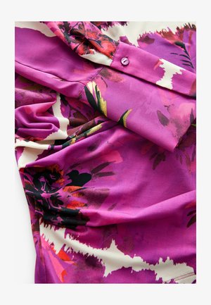 Blouse violette avec un motif floral comportant des accents roses, rouges et noirs. Fabriquée en tissu doux, elle inclut un détail de bouton au niveau du poignet.