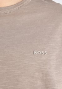 Tricou bej deschis, dintr-un material moale, cu guler rotund și un mic logo "BOSS" în ton cu materialul, amplasat în zona stângă a pieptului.