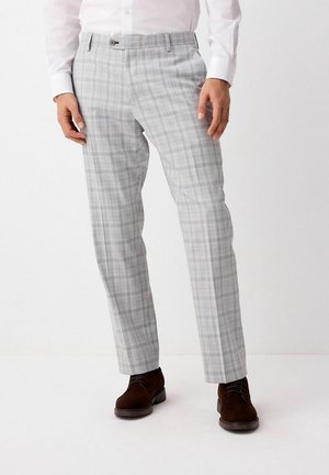 Mężczyzna ubrany w szare spodnie w kratę, białą koszulę na guziki oraz ciemnobrązowe sznurowane buty, stojący na tle jednolitej białej ściany.