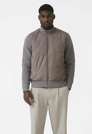 Homme portant un gilet matelassé taupe par-dessus un pull côtelé gris avec un pantalon beige clair, debout sur un fond blanc uni.