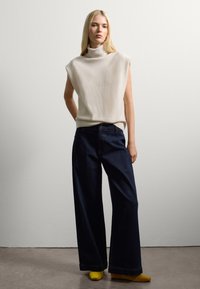 Helle beige ärmellose Rollkragenpullover, kombiniert mit dunkelblauen weiten Jeans und gelben Schuhen. Einfache Textur und lockere Passform.