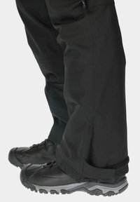 Black waterproof pants with a tapered design, featuring a Velcro adjustment at the ankle and worn with rugged black shoes.
