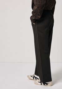 Pantaloni neri a gamba larga con una trama liscia, caratterizzati da strisce laterali marroni e tasche sui lati, abbinati a sneakers bianche e nere.