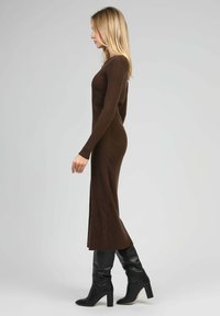 Robe en tricot côtelé marron avec des manches longues, silhouette ajustée, fente sur le côté, portée avec des bottes noires à talons hauts jusqu'aux genoux. Texture lisse.