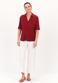 Camisa roja de manga corta con cuello, dos bolsillos en el pecho y parte delantera abotonada. Combinada con pantalones de pierna ancha blancos y zapatos planos marrones.