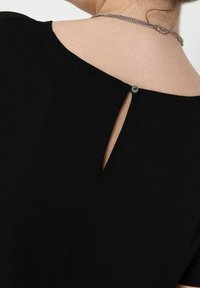 Vue arrière d'une personne portant un haut noir avec une petite ouverture en clé boutonnée près de l'encolure et un collier en chaîne argentée.