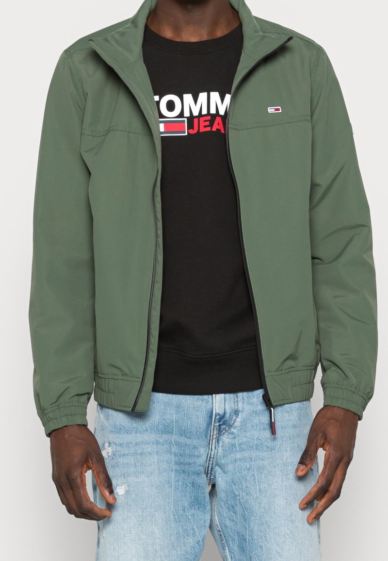 Muž v nezapnuté olivově zelené bundě přes černou mikinu Tommy Jeans a světle modrých roztrhaných džínách, stojící s uvolněnými pažemi.