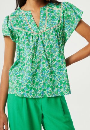 Femme portant un chemisier à manches courtes à motif floral vert et bleu avec bordure en dentelle et un pantalon vert uni, debout avec une main sur la hanche.