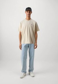 Beiges Baumwoll-T-Shirt mit kleinem Logo auf der linken Brust, kombiniert mit hellblauen Jeans mit geradem Bein und weißen Sneakern. Schwarze Kappe inklusive.