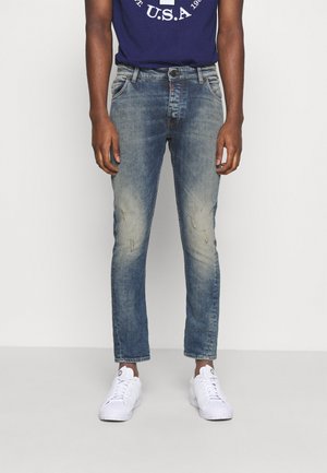 Hombre vestido con unos jeans ajustados azul desgastados, zapatillas blancas y una camiseta azul marino con un estampado gráfico blanco.