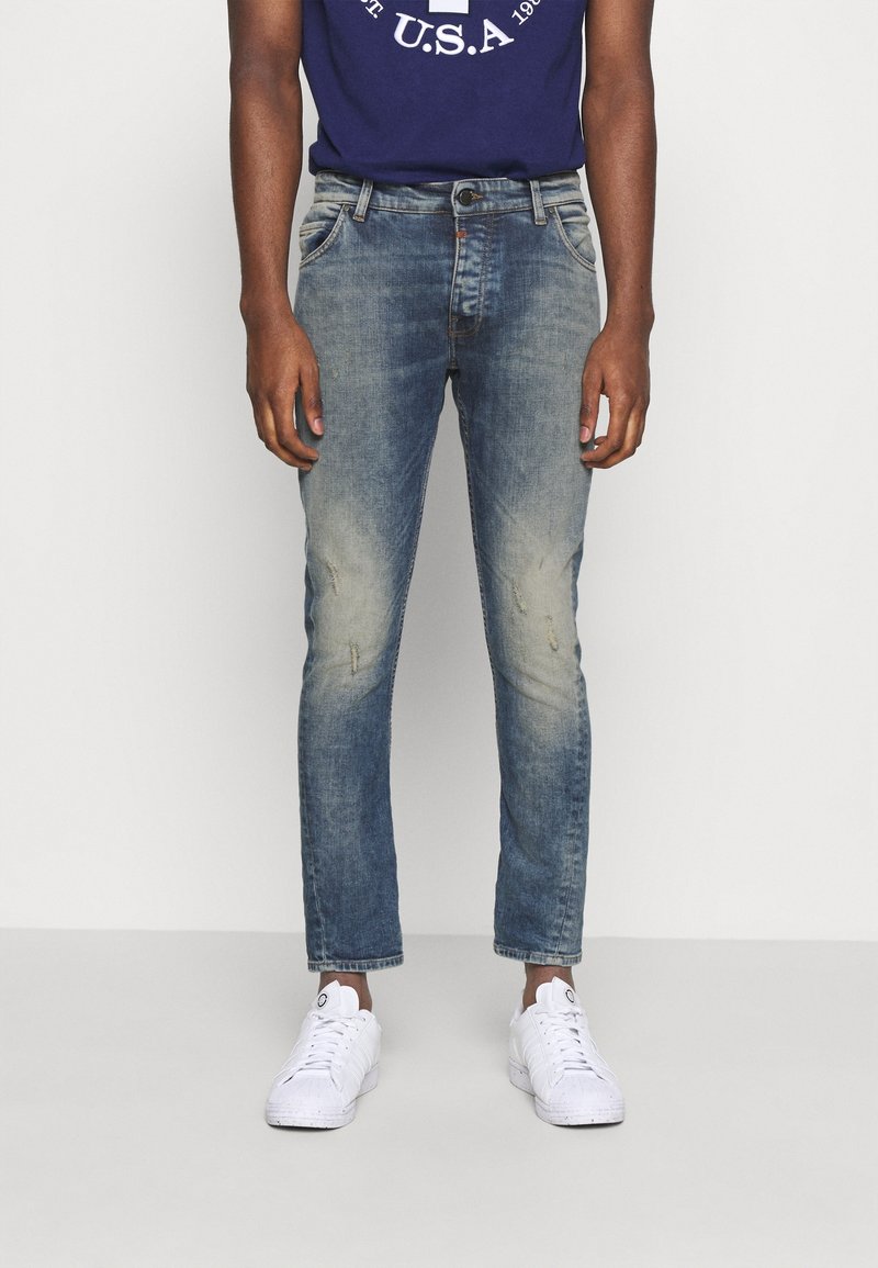 Mężczyzna w wyblakłych, dopasowanych niebieskich dżinsach, białych sneakersach i granatowej koszulce z białym nadrukiem.