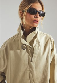 Bež jakna oversize z visokim ovratnikom in kovinskim obročem. Model nosi črne pravokotne sončne očala, ki predstavljajo čist in moderen dizajn.