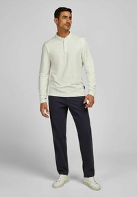 Weißes Langarm-Henley-Shirt mit einem geknöpften Kragen, kombiniert mit dunkeln Marineblau-Hosen und weißen Sneakern, vor einem neutralen Hintergrund.