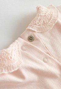 Prenda de algodón rosa pálido con cuello adornado con encaje, que cuenta con dos cierres de presión. Textura suave y tejido delicado.