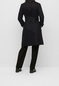 Cappotto nero dal taglio su misura, con colletto alto e una texture morbida. Indossato sopra pantaloni neri larghi con strisce laterali.