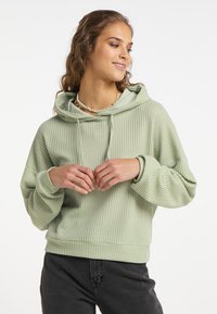 Felpa verde chiaro con texture, dal taglio rilassato e cappuccio con coulisse regolabile, abbinata a pantaloni grigio scuro e una collana di perline.