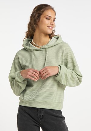 Felpa verde chiaro con texture, dal taglio rilassato e cappuccio con coulisse regolabile, abbinata a pantaloni grigio scuro e una collana di perline.