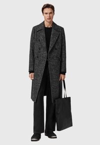 Sort og grå tekstureret frakke med dobbeltradede knapper, båret over en sort striktrøje og mørke bukser, parret med en sort tote bag.