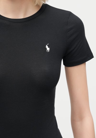 Nærbilde av en person som har på seg en tettsittende svart t-skjorte med korte ermer og en liten hvit brodert logo av en polo-spiller på brystet.