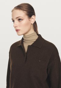 Bruine gebreide poloshirt met een kraag, featuring een knopdetail en een klein borstzakje, gecombineerd met een beige turtleneck.