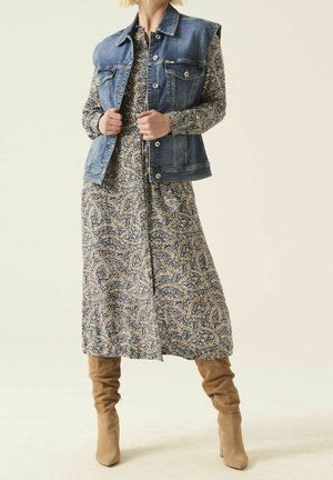 Nainen pukeutuneena kuvioituun pitkähihaiseen midimekkoon, hihattomaan siniseen farkkutakkiin ja vaaleanruskeisiin mokkanahkaisiin polvenkorkuihin saappaisiin.