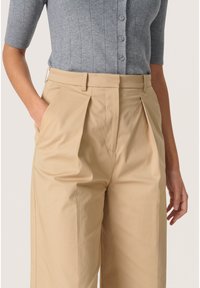 Pantaloni plisati bej, cu buzunare laterale si o textura neteda, asortati cu un cardigan gri deschis cu dungi, prevazut cu nasturi pe fata.