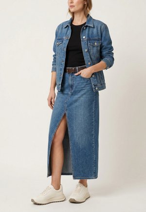 Frau trägt eine blaue Jeansjacke und einen passenden langen Jeansrock mit Frontschlitz, schwarzes Shirt, brauner Gürtel und weiße Sneakers.