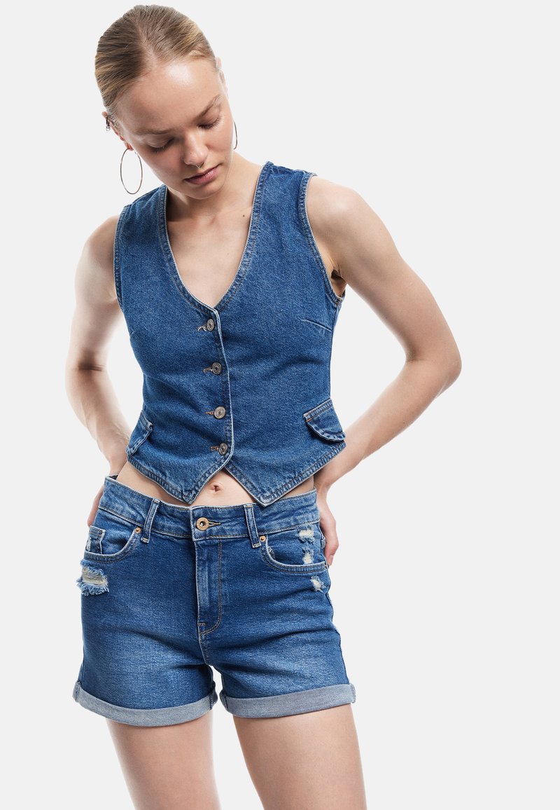 Mujer vestida con un chaleco de mezclilla sin mangas y pantalones cortos de mezclilla desgastados, mirando hacia abajo con las manos reposando en las caderas.