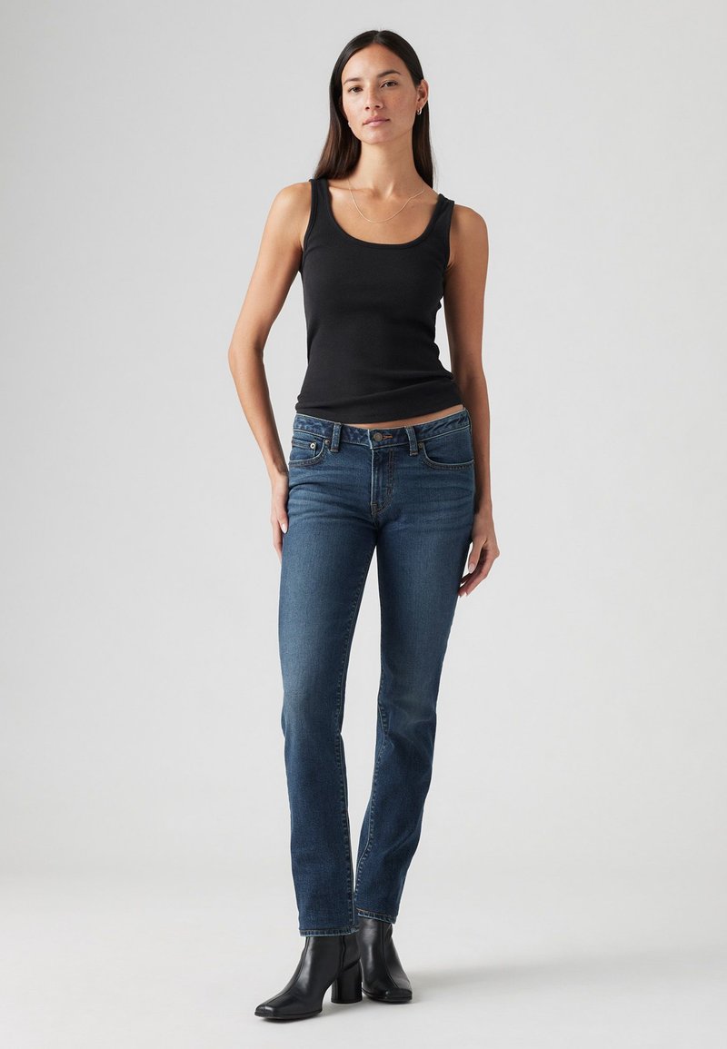 Top nera senza maniche, jeans blu con una leggera sfumatura, cuciture chiare e forma aderente; stivali neri alla caviglia con tacco alto completano l'outfit.