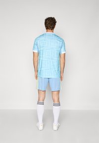 Hellblaues Sporttrikot mit einem gemusterten Design, kombiniert mit passenden Shorts. Mit weißen Akzenten und drei schwarzen Streifen auf knielangen Socken.
