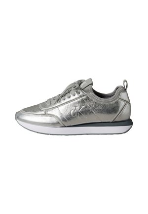 Zapatilla deportiva de perfil bajo plateada metálica con cordones, suela negra y blanca, y logo "cK" en el lateral, vista desde el lado exterior.