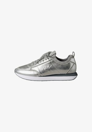 Zapatilla deportiva de perfil bajo plateada metálica con cordones, suela negra y blanca, y logo "cK" en el lateral, vista desde el lado exterior.