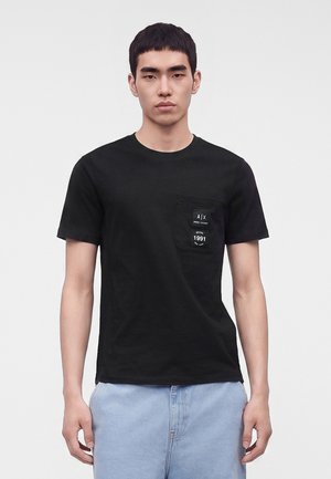Jeune homme portant un t-shirt noir avec une poche poitrine et un jean bleu clair, debout devant un fond gris clair uni.