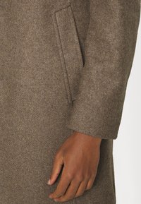 Manche de manteau en laine marron avec une poche fendue sur le côté, une main à la peau foncée reposant à côté, sur un fond uni.
