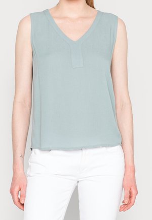 Vrouw draagt een mouwloos lichtblauw V-hals topje en een witte broek, staand tegen een effen lichte achtergrond.