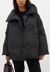 Crna puffer jakna s visokim ovratnikom, oversized kroja i vidljivim dugmadima. Glatka tekstura, s minimalističkim dizajnom.
