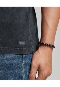 Camiseta de algodón gris oscuro con una pequeña etiqueta en el dobladillo, combinada con jeans de mezclilla y una pulsera de cuentas de madera oscura en la muñeca.