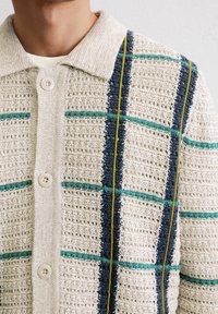 Beige Strickjacke mit strukturiertem Gewebe, versehen mit einem karierten Muster in Blau, Petrol und Gelb, zwei Knopfverschluss und einem Kragen.
