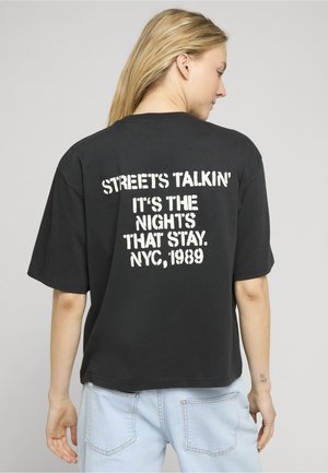 Femme aux longs cheveux blonds portant un t-shirt noir avec un texte blanc "STREETS TALKIN' IT'S THE NIGHTS THAT STAY. NYC, 1989" et un jean bleu clair.