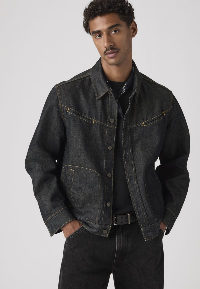 Mand iført en sort denimjakke med gule syninger, sorte bukser og hænderne i lommerne mod en ensfarvet grå baggrund.