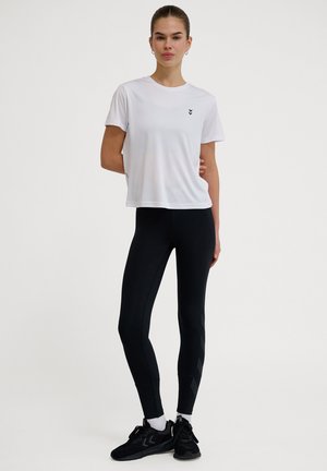 Hvid trænings t-shirt med korte ærmer og sort logo; sorte leggings med teksturerede sidelommer; sorte træningssko med snørebånd.