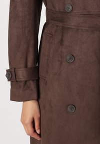 Brązowy zamszowy płaszcz trench z paskiem w talii, dużymi guzikami i miękką teksturą, zbliżenie na mankiet i detale.