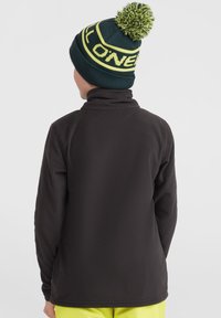Pull en polaire noir avec un col haut, porté avec un bonnet vert foncé orné d'un pompon jaune et d'accents textuels. Vu de dos.