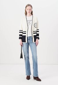 Cardigan crem cu dungi bleumarin, tricou alb cu grafică, blugi deschiși la culoare evazați, geantă neagră și cizme maro cu toc. Textură netedă.