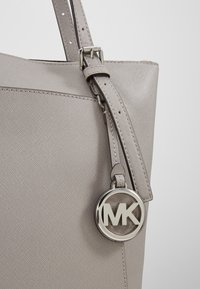Grijze, gestructureerde stoffen tas met een textuur, voorzien van een zilveren MK-logo hanger die aan de hengsels is bevestigd en een glanzende afwerking op het materiaal.