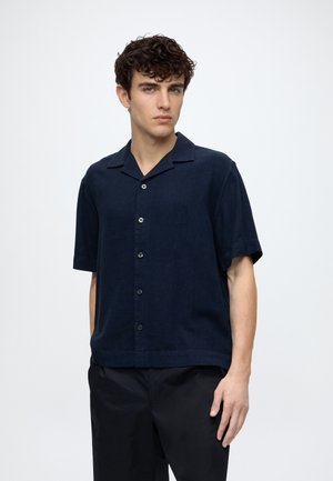 Jeune homme aux cheveux bouclés portant une chemise bleu marine foncé à manches courtes boutonnée et un pantalon noir sur un fond blanc uni.